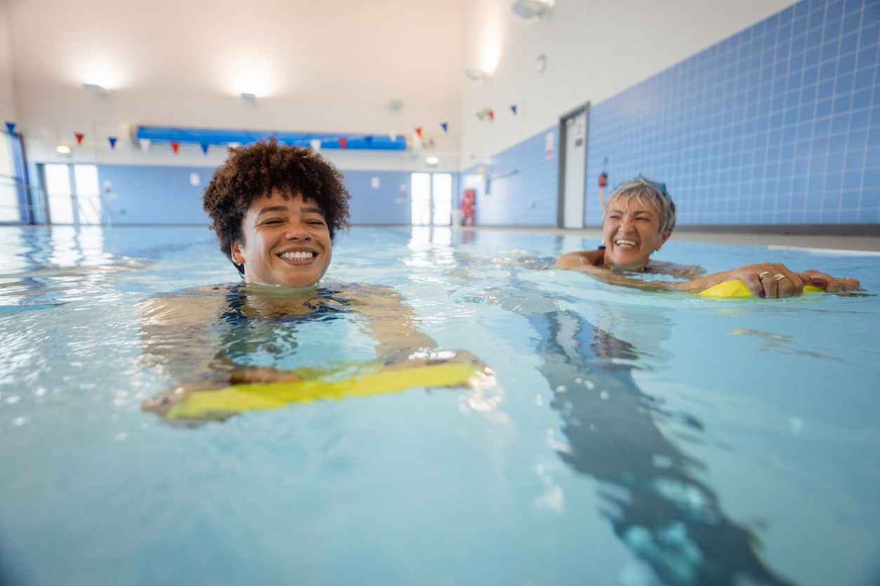A senior women swimming while using a float alongside a swimming instructor in a swimming pool in Boldon, North East England. One woman is looking at the other and smiling, health benefits of swimming